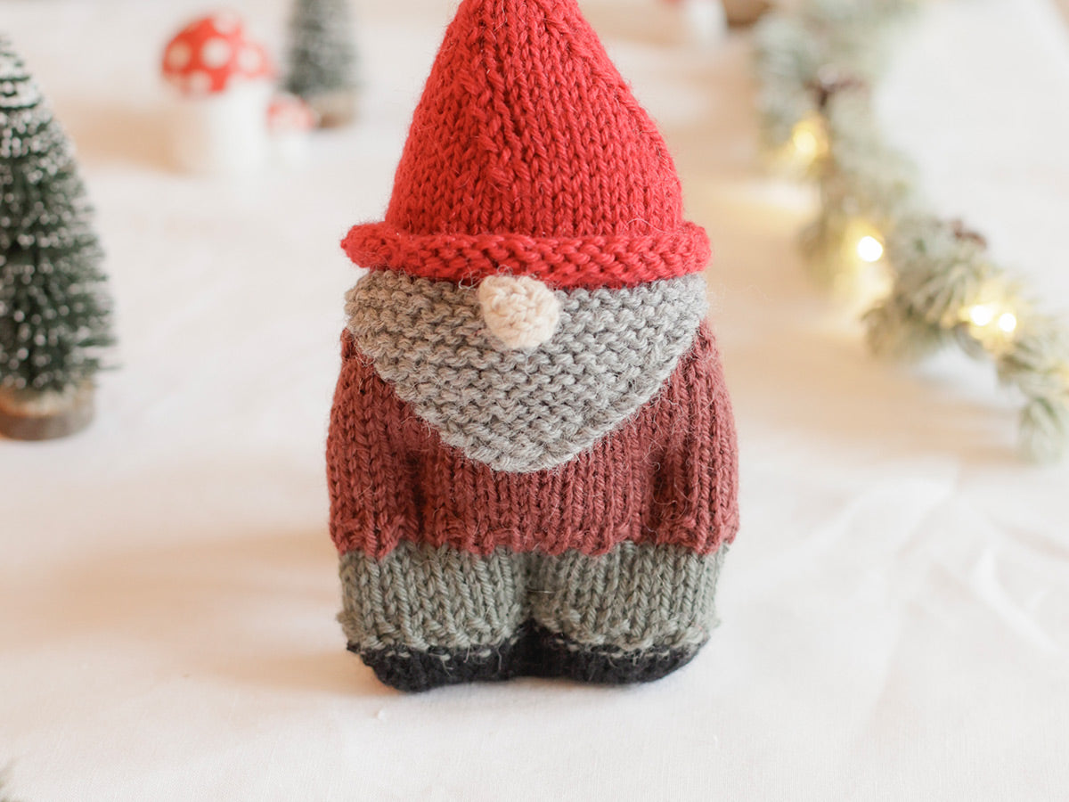 Knitted gnome with a red hat and brown outfit on a soft surface with blurred Christmas trees in the background.